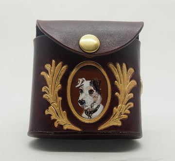 Mahogany leather dog waste bag dispenser with brass snap hardware. Design features a portrait of a jack russell terrier dog against an umber colored background with a gold frame, wreathed with gold laurels.