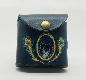 Peacock teal leather dog waste bag dispenser with brass snap hardware. Design features a portrait of a pomeranian dog against a monastral blue colored background with a gold frame, wreathed with gold peacock feathers.