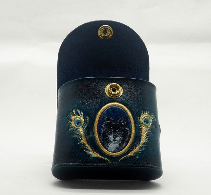 Peacock teal leather dog waste bag dispenser with brass snap hardware, photographed with the snap opened to showcase the navy blue interior. Design features a portrait of a pomeranian dog against a monastral blue colored background with a gold frame, wreathed with gold peacock feathers.
