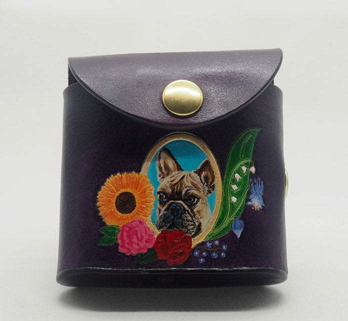 Eggplant purple leather dog waste bag dispenser with brass snap hardware. Design features a portrait of a french bulldog against a bright blue colored background with a gold frame, wreathed with an arrangement of flowers.