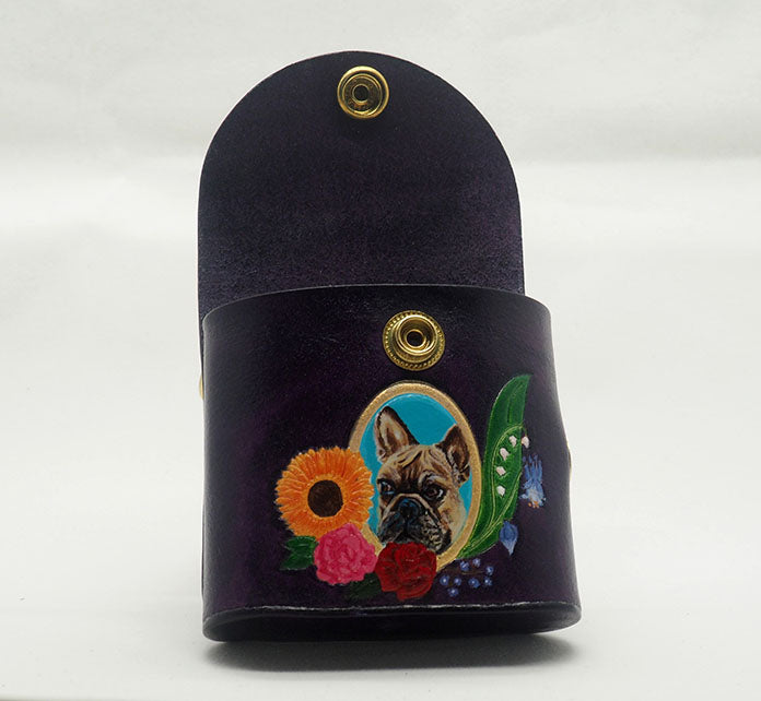 Eggplant purple leather dog waste bag dispenser with brass snap hardware. Photographed with the snap open to showcase the interior color is the same as the exterior. Design features a portrait of a french bulldog against a bright blue colored background with a gold frame, wreathed with an arrangement of flowers.